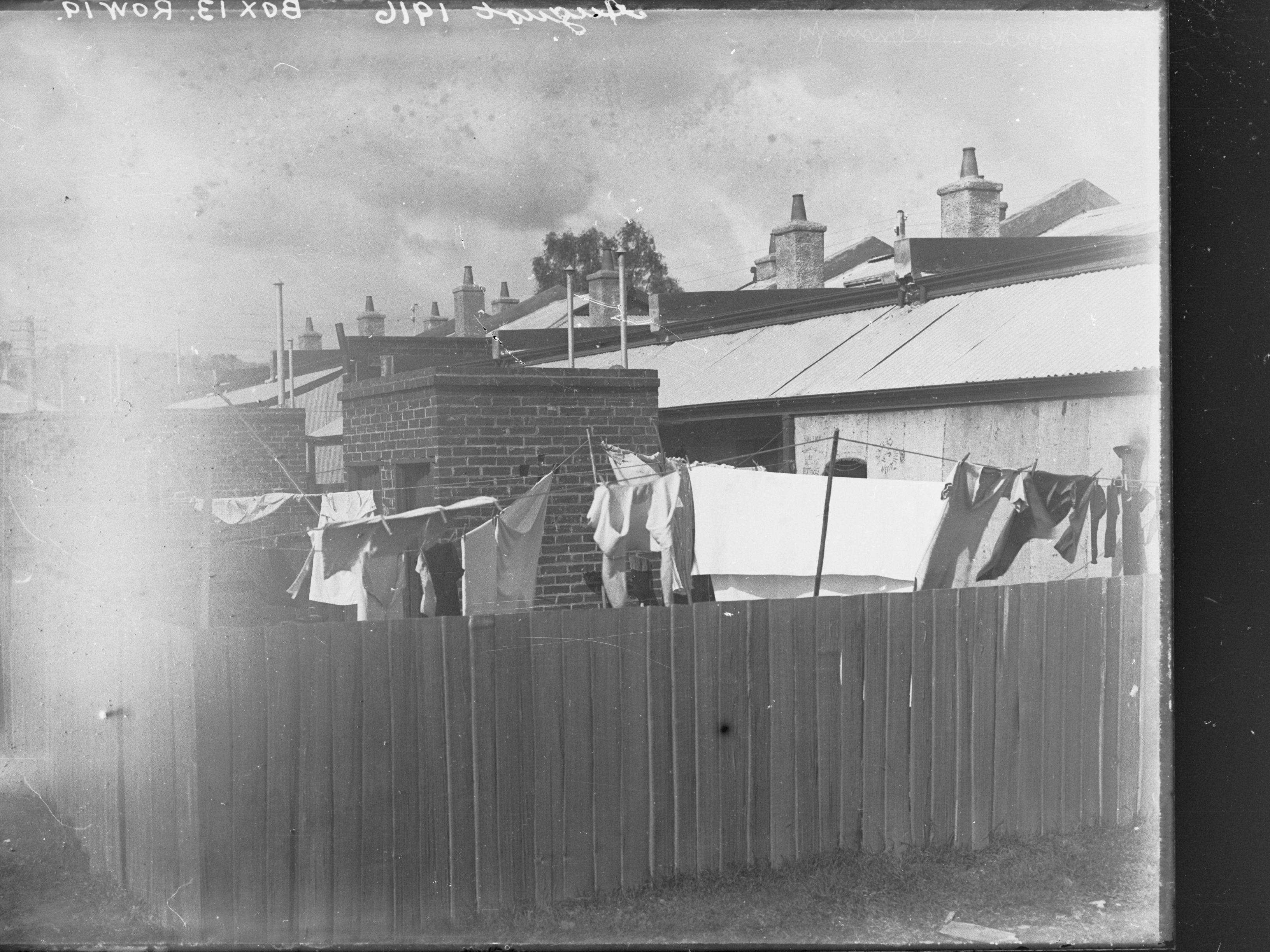 A black and white photograph of a fenced-in backyard. There are clothes hanging on a clothes line, visible behind the fence.